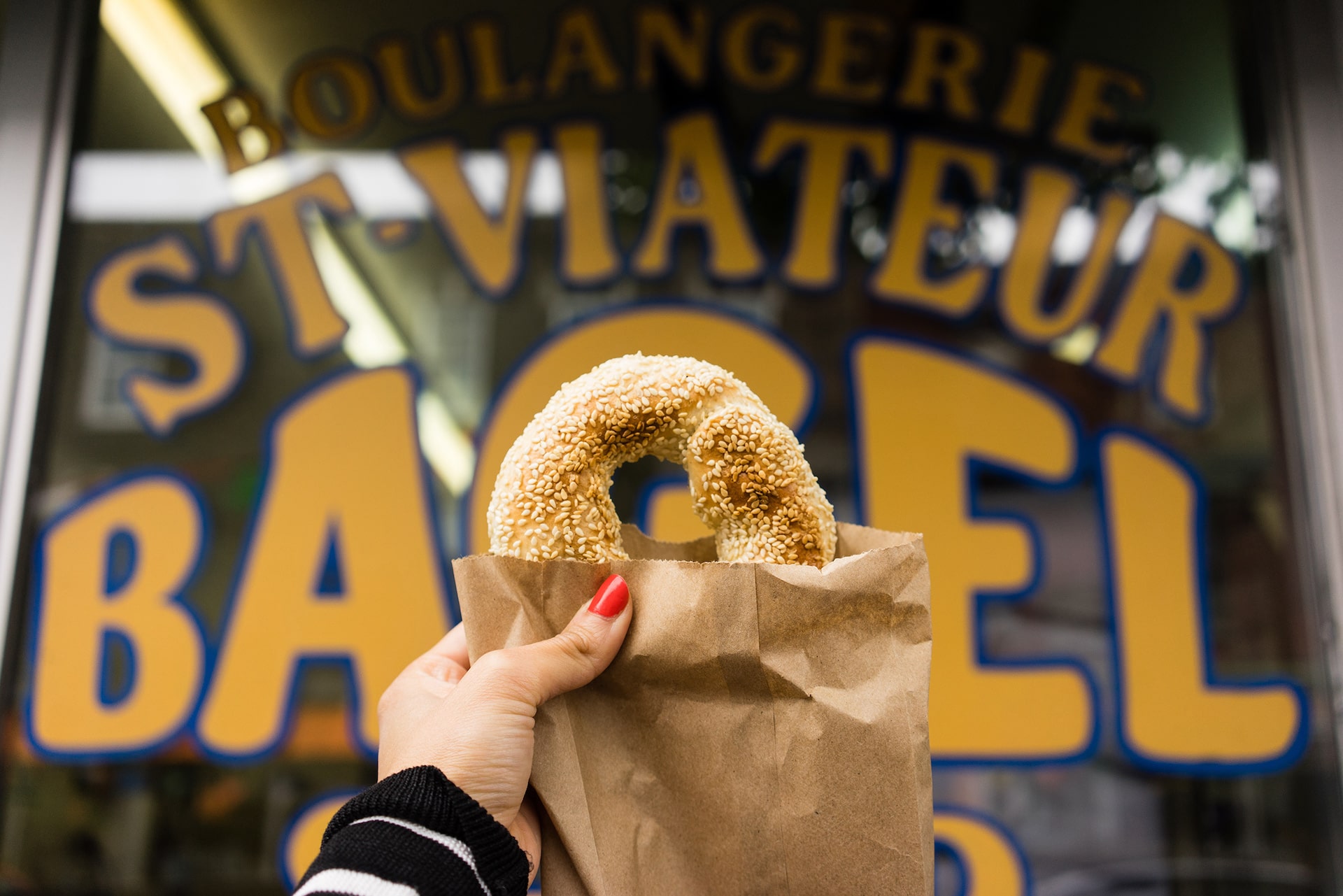 Gros plan sur un Bagel tenu par une main de femme devant une vitrine d'un magasin de bagels